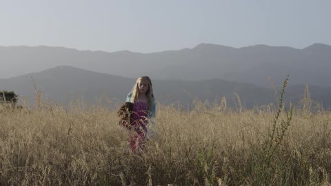 A young blonde girl carrying a doll and walking through tall grass with mountains in the background