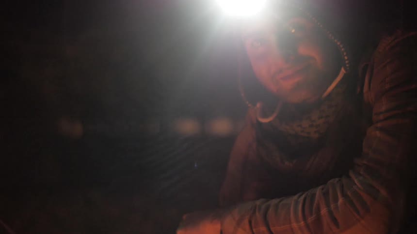 A man wearing a miner's light on his head adjusting something on the ground and then looking towards the camera while smiling and speaking