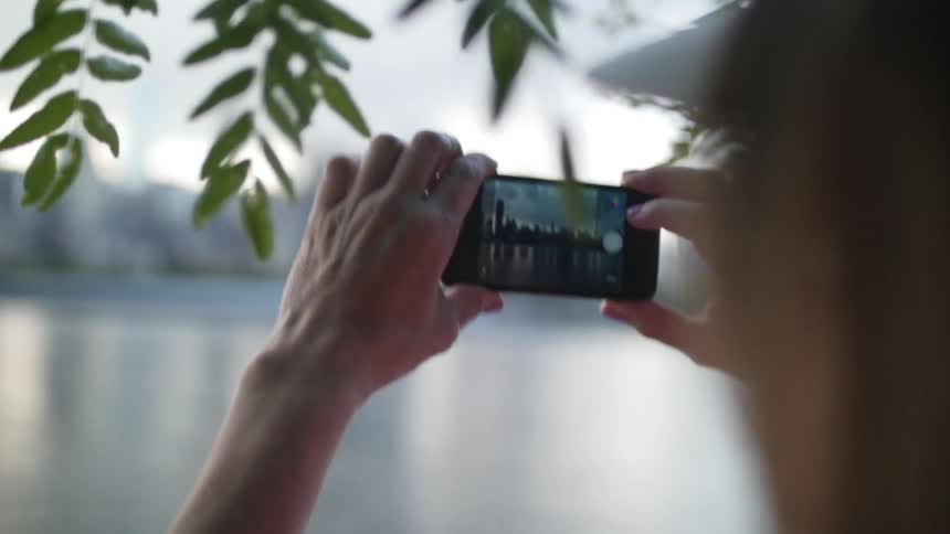 A woman takes a cellphone picture of a city across a body of water