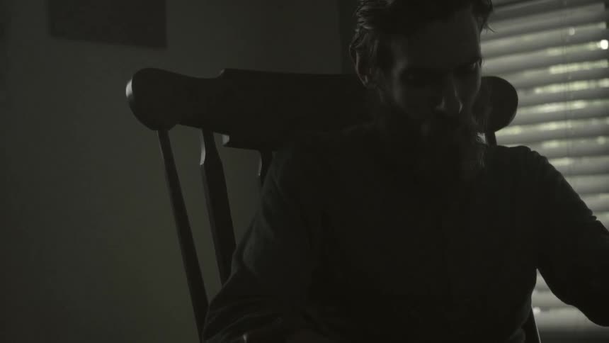 Man sitting in a chair reading a book near a window in the dark