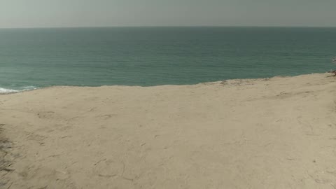 First person view of walking to the edge of a sandy cliff and looking down at the rocky shoreline as waves crash on shore