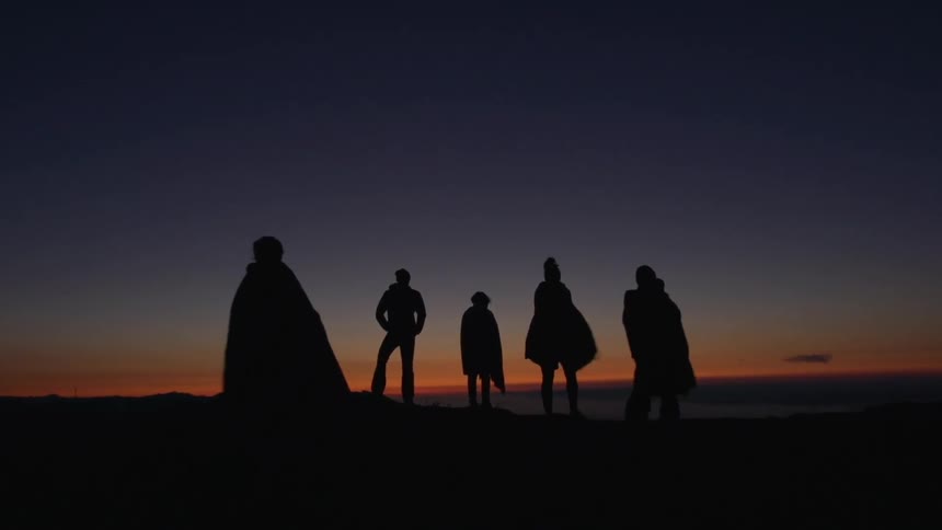 Four people standing at a cliff edge while three others run up to join them during sunset