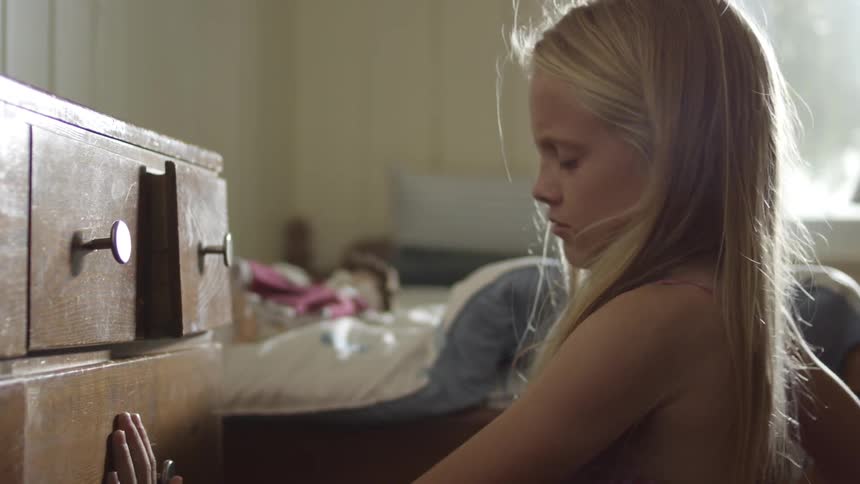 A blonde girl pulls clothing from a dresser