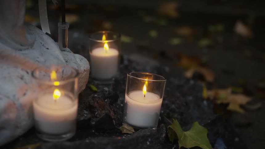 Lit candles in glass containers are on the ground with scattered fall leaves