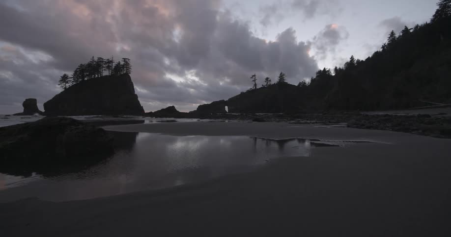 The sandy shore of a body of water surrounded by cliffs and trees during a very cloudy sunset