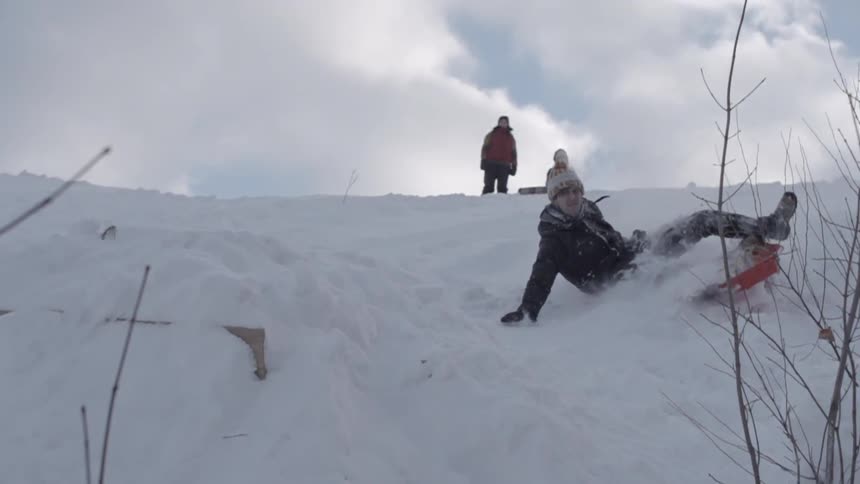 Children sitting on top of a snow-covered hill as another child slides down the hill kneeling on a sled before falling off