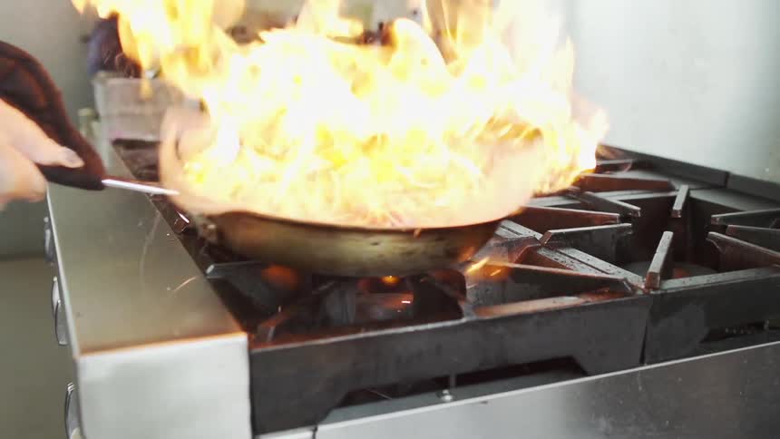 A chef flames a saute pan full of onions over a commercial stove in a kitchen