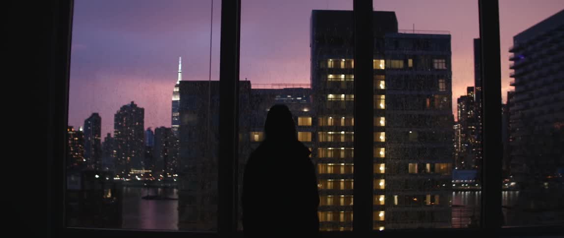 A person standing in front of a window as lightning flashes in sunset sky