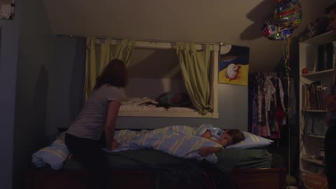 A woman helping a young girl climb over another girl so she can get into bed and then kissing another girl before smoothing her hair