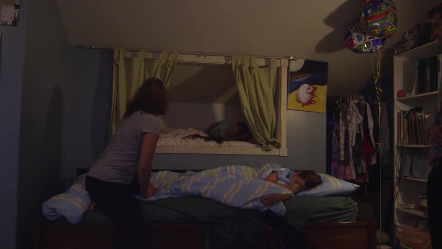 A woman helping a young girl climb over another girl so she can get into bed and then kissing another girl before smoothing her hair