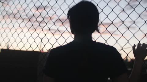 A young woman clinging onto chain link fencing a looking out over an urban area