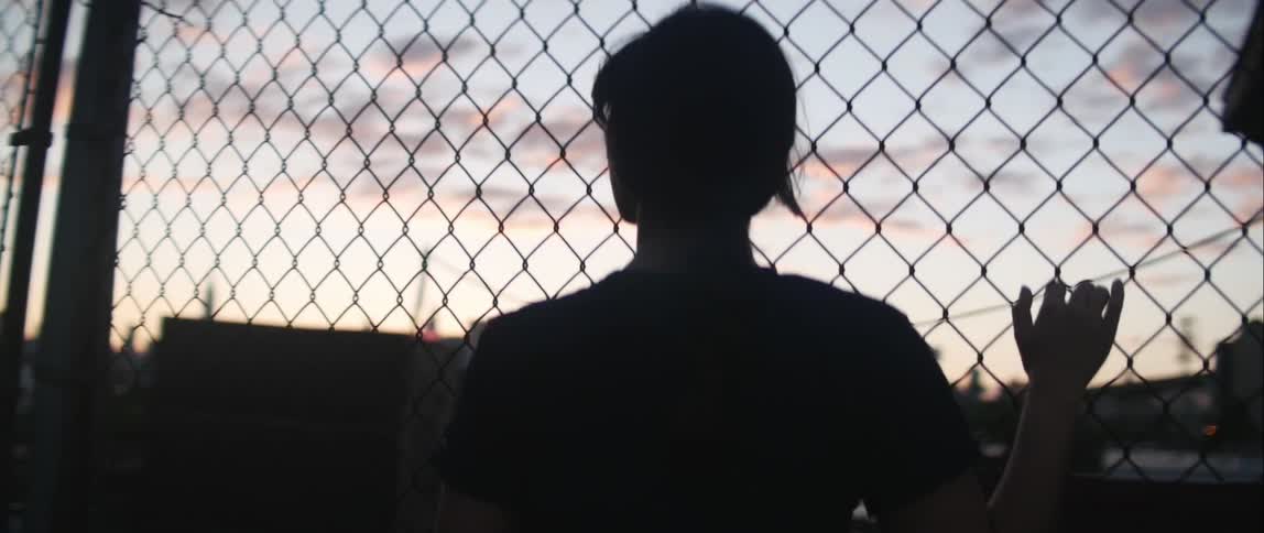 A young woman clinging onto chain link fencing a looking out over an urban area