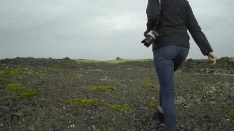 A woman carrying a camera and walking along rocky muddy ground on a cloudy day