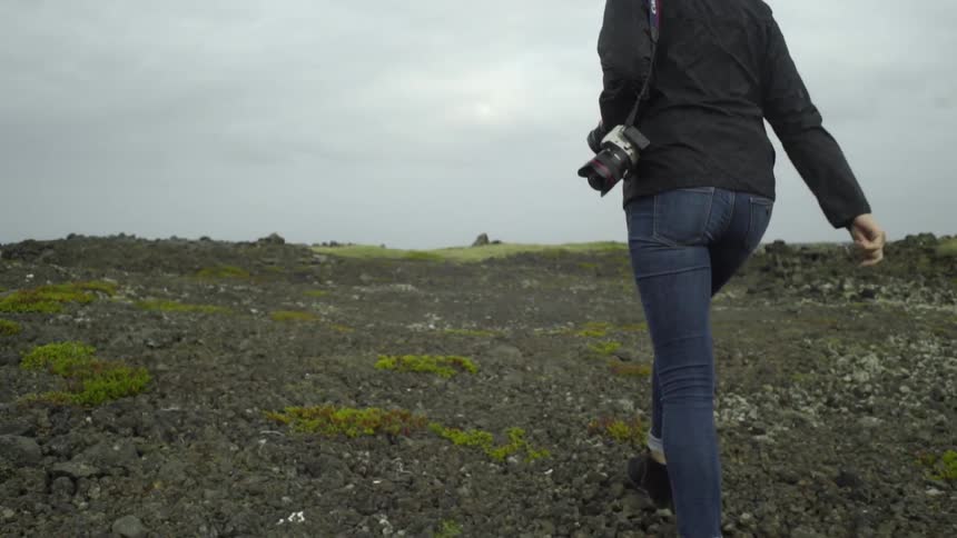 A woman carrying a camera and walking along rocky muddy ground on a cloudy day