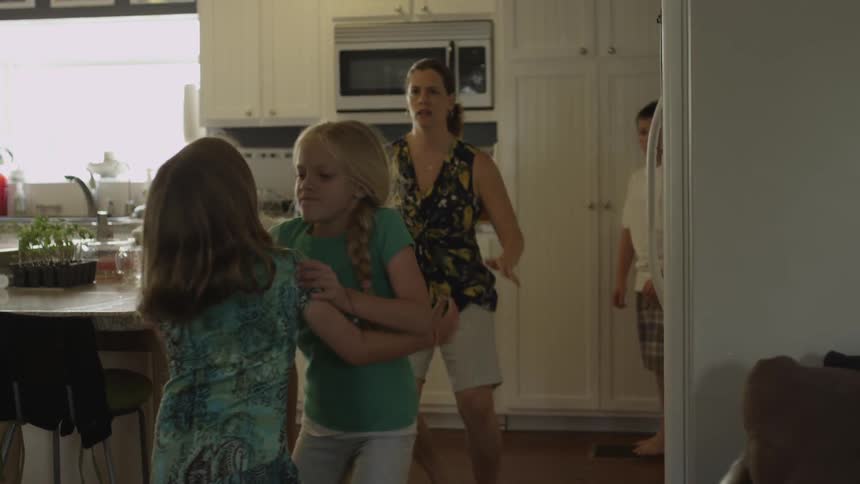 A woman trying to hold a young blonde girl back before she breaks away and shoves another young girl down in a kitchen