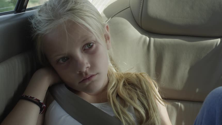 A young blonde girl riding in a car and looking sad