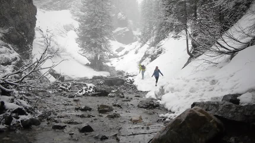 Two people walk along a snow covered river bank as snow falls