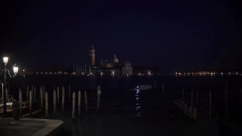 A boat cruising away from docks at night while lights shine from buildings in the background