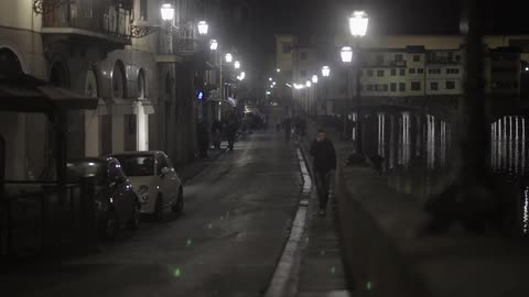 Several people walk down a narrow street under the streetlamps at night, while one of the people appears to be talking on his cellphone
