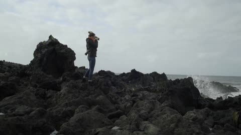 A young woman in tight jeans stands on the rocks of a beach taking photographs of the crashing waves