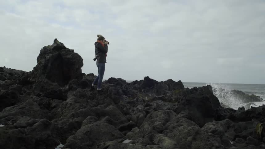 A young woman in tight jeans stands on the rocks of a beach taking photographs of the crashing waves