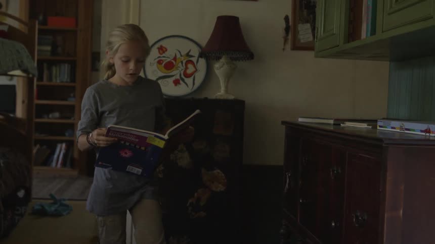 A young blonde girl walking through a doorway with an open book when she stops to listen to something