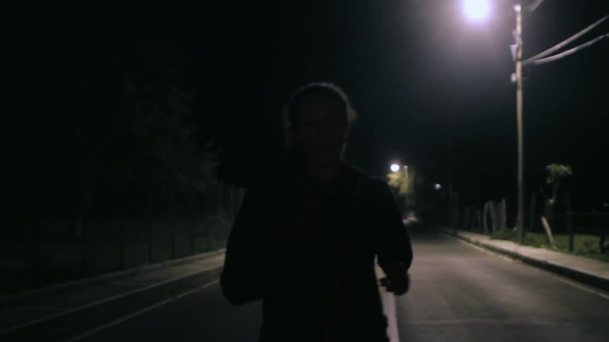 A person with their hair in a ponytail jogs down a deserted street under the street lights at night