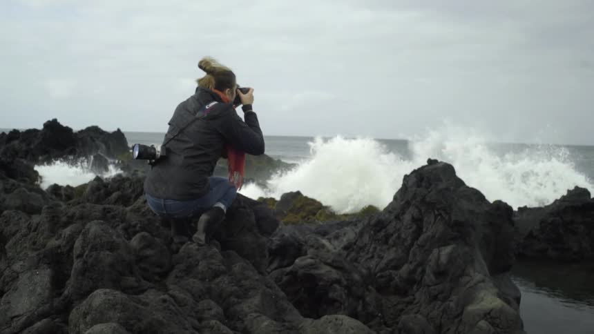 A woman crouches on boulders and photographs the turbulent ocean