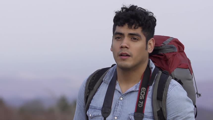 A young man wearing a large backpack and walking with mountains in the background
