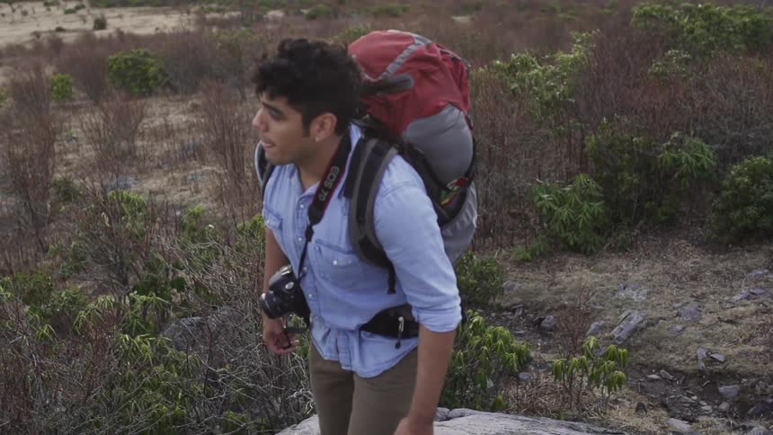 A man wearing a backpack and a camera around his neck while climbing a boulder