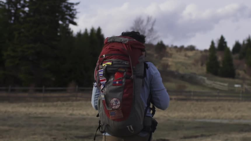 A man wearing a backpack hikes across a field bounded by a wooden fence