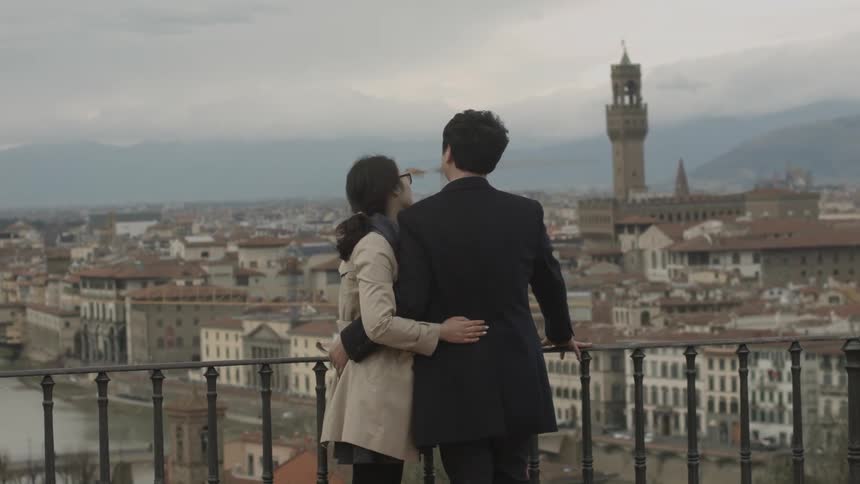 A couple embrace as they stand on a balcony that overlooks a city neighborhood