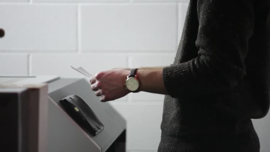 A man sitting with is hands on his knees before jumping up and grabbing a piece of paper out of a receptacle and reading it