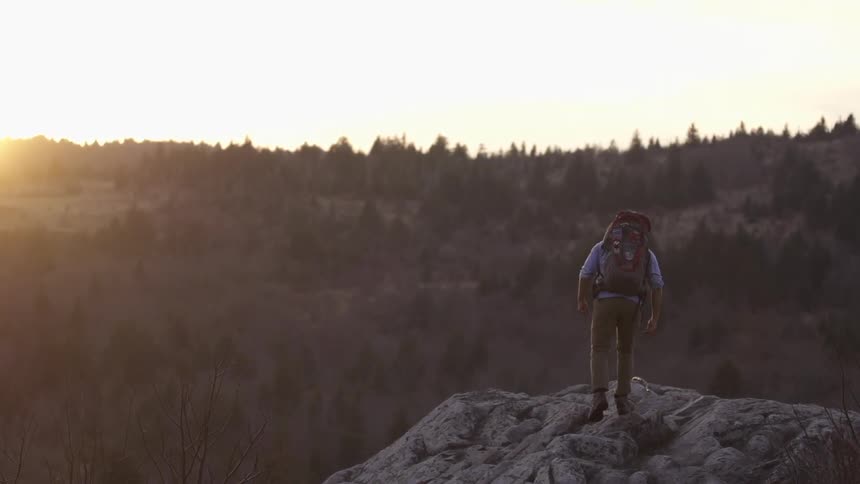 A young man wearing a large backpack and hiking to the top of a boulder to look out over the mountains