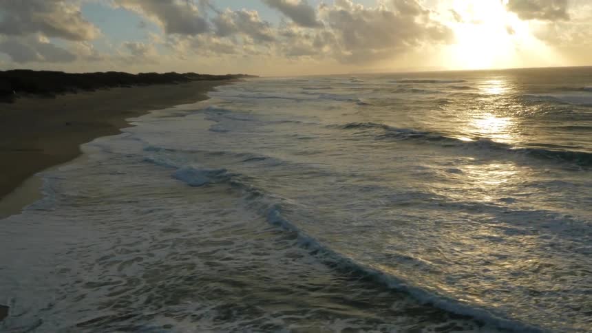 Waves crashing onto shore line as people stand on the beach at sunset