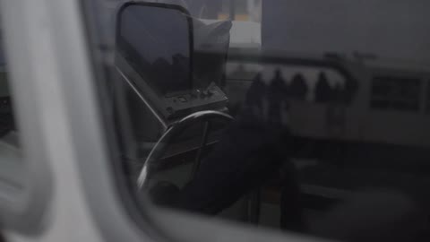 Someone turning the steering wheel on a boat as the reflection of a passenger ferry is seen in the window