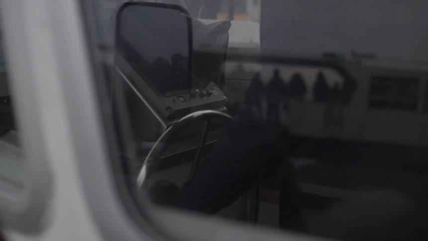 Someone turning the steering wheel on a boat as the reflection of a passenger ferry is seen in the window