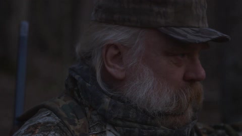 An older man with long hair and a beard wearing hunting gear outside at night
