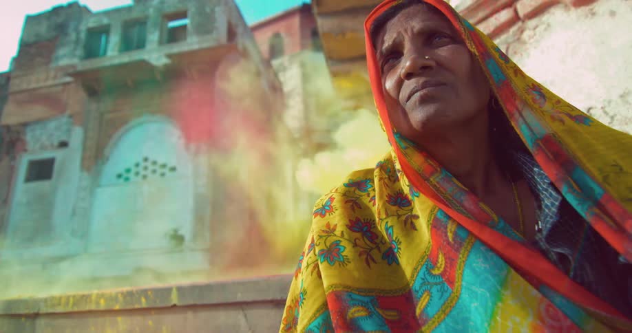 A woman wears a colorful scarf covering her head as she sits outside and colorful chalk dust flies behind her