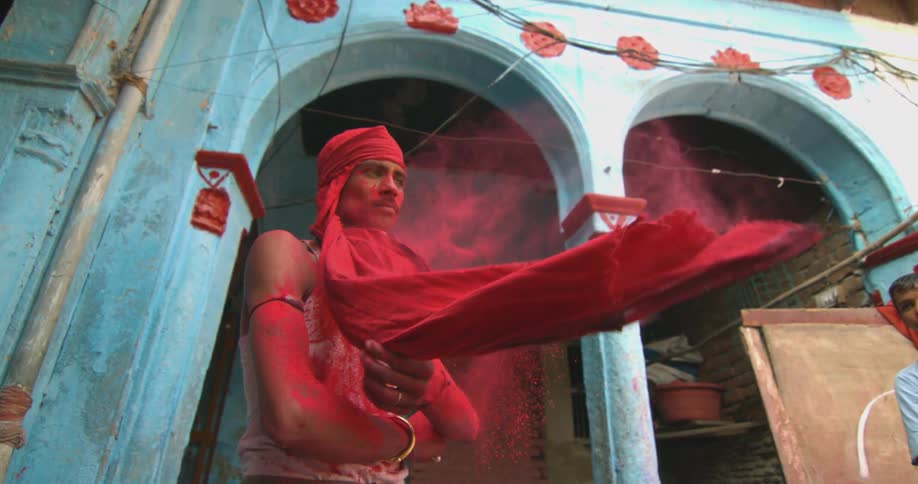 Man covered in red powder removes a cloth from his face