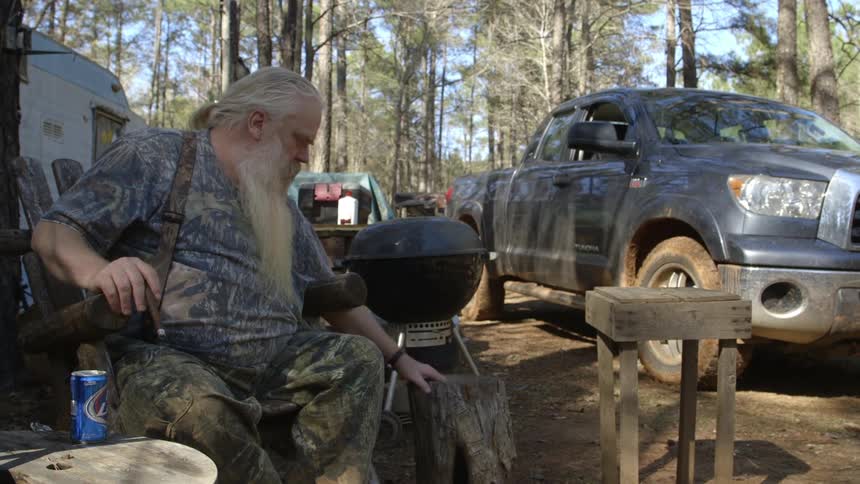 A heavy-set, bearded man sitting next to a grill, smoking a cigar and drinking a beer at a campsite