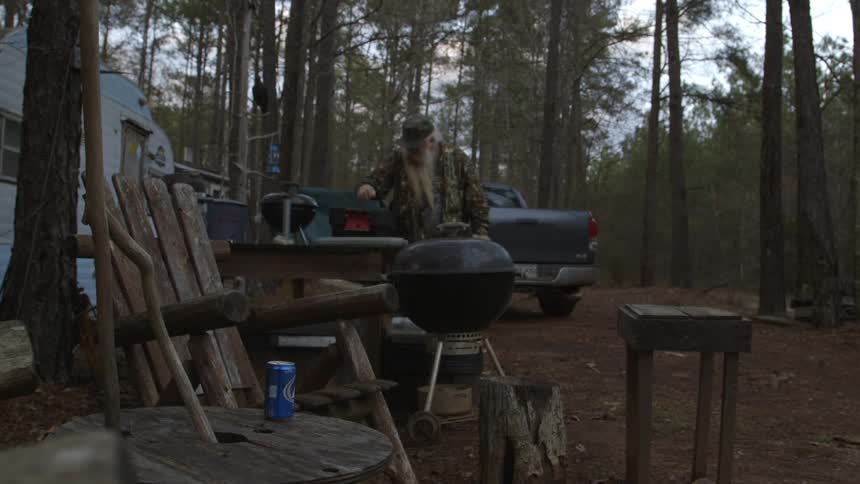 A man carries a box to a table outside of a camper in the woods