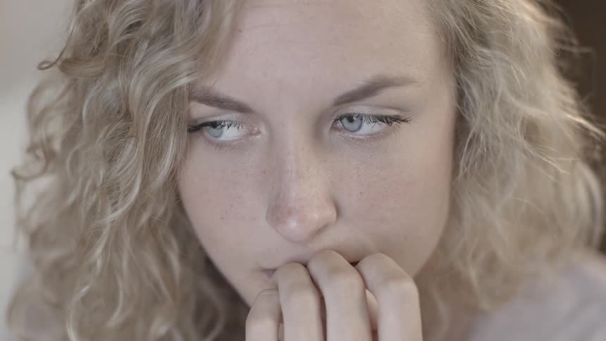 A blonde woman chews on her nails intently and then inspects her nail to see if she got the rough spot