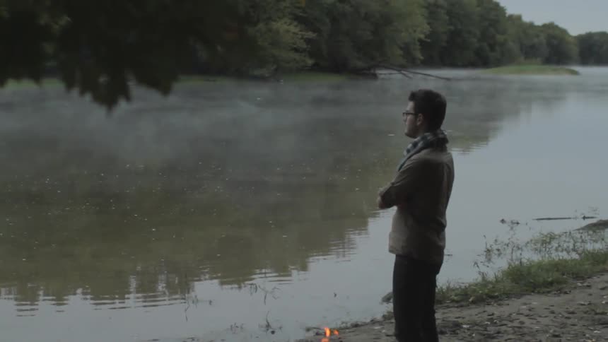 A man stands next to a campfire by a river
