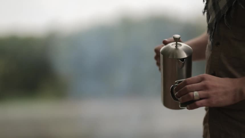 A man standing outdoors pours hot coffee from a metallic pitcher into a coffee cup