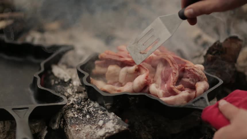 A person cooks bacon in a skillet over an open fire