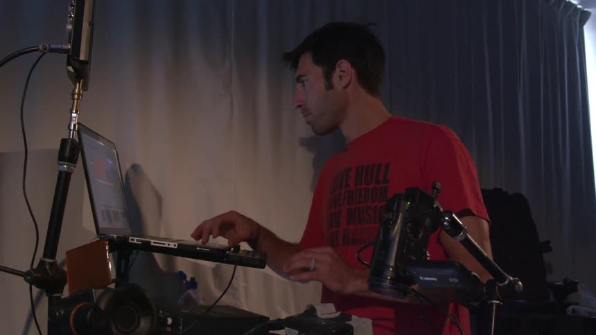 A brunette man typing on a laptop while sitting behind audio equipment