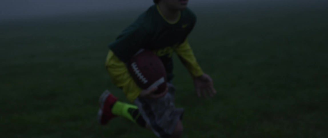 A young boy wearing yellow shorts handing a football off to a young boy wearing a short-sleeved green shirt over a long-sleeved yellow shirt on a foggy day