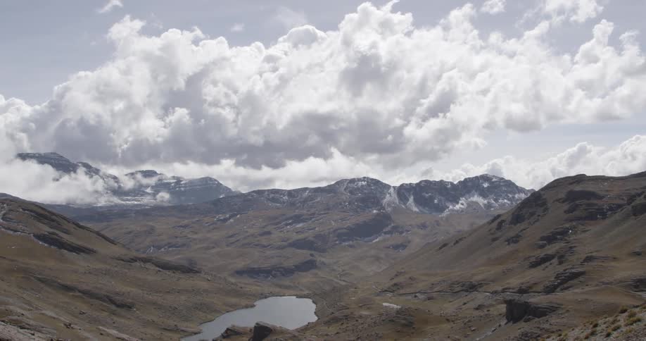 Many fluffy white clouds in the sky above a grassy mountain area with a body of water