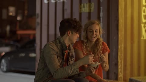A young playful couple sit on an outside bench sharing a bowl of food